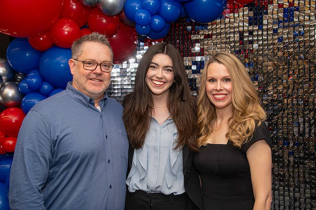 Three people are smiling in front of a colorful balloon and mirrored background.